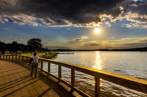 A person walks along a wooden path by a lake at sunset, with dramatic clouds and sunlight reflecting on the water.