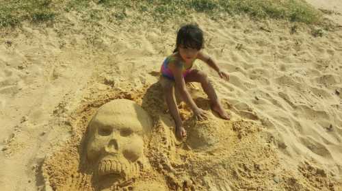 A child plays beside a large sand sculpture of a skull on a beach.