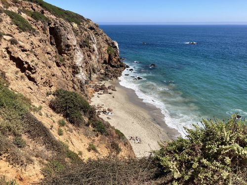 A scenic view of a rocky coastline with a sandy beach and calm blue ocean under a clear sky.