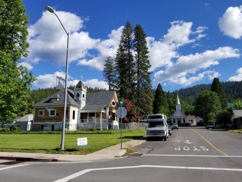A quaint street scene featuring a large house, a church, and tall trees under a blue sky with fluffy clouds.