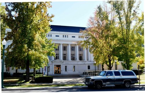 A classic building with columns surrounded by trees, and a parked truck in front on a sunny day.