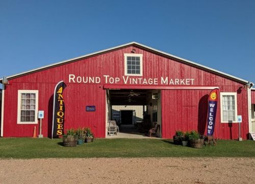 Red barn exterior of Round Top Vintage Market with banners for antiques and a welcome sign. Clear blue sky above.