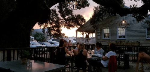 A group of people dining outdoors at sunset, with trees and a cozy restaurant in the background.