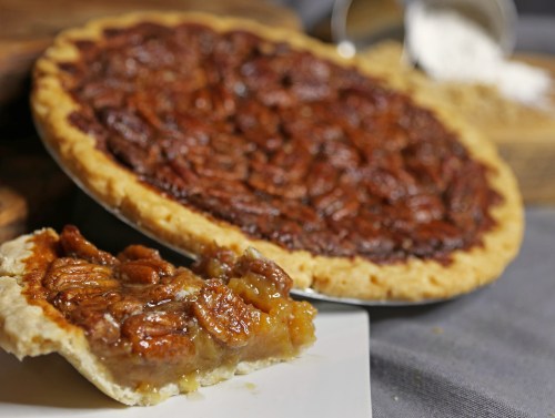 A slice of pecan pie on a plate, with the whole pie in the background, showcasing a golden crust and rich filling.