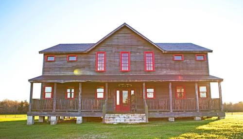 A rustic wooden house with red accents, featuring a spacious porch and large windows, set against a bright sky.