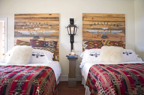 Cozy bedroom with two beds, rustic wooden headboards, colorful blankets, and decorative pillows. Warm, inviting atmosphere.