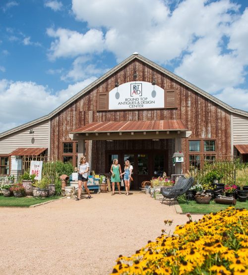 Three women walk outside a rustic building with a sign for an antique center, surrounded by flowers and greenery.
