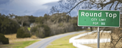 Green city limit sign for Round Top, Texas, indicating a population of 90, with a rural road in the background.