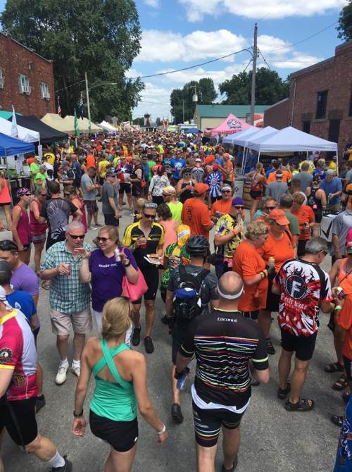 A vibrant crowd of people in colorful attire gathers at an outdoor event, with tents and trees in the background.