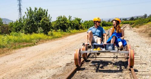 A couple in helmets sits on a rail cart on a sunny day, surrounded by greenery and a dirt path.