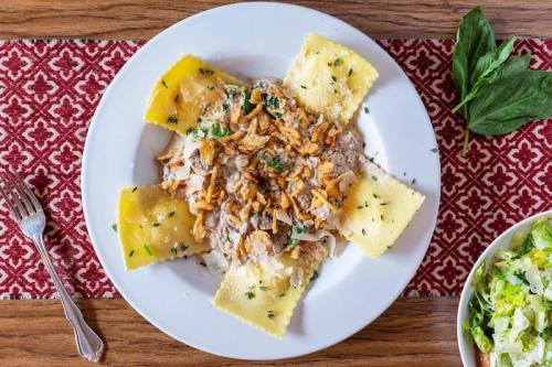 A plate of ravioli topped with onions and crispy shallots, garnished with herbs, alongside a side salad.