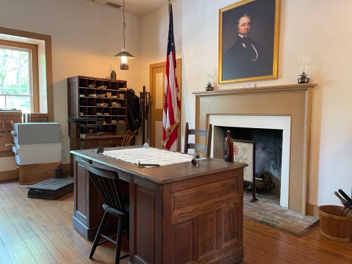 A historic office with a wooden desk, chair, American flag, and a portrait on the wall, featuring a fireplace and shelves.