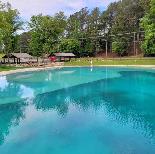 A clear blue swimming pool surrounded by green trees and picnic pavilions on a sunny day.