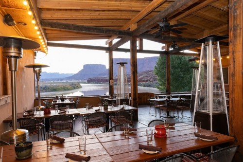 A rustic restaurant patio with wooden tables, heaters, and a scenic view of mountains and a river.
