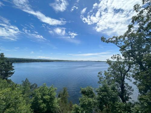 A serene lake view surrounded by lush greenery and a clear blue sky with scattered clouds.