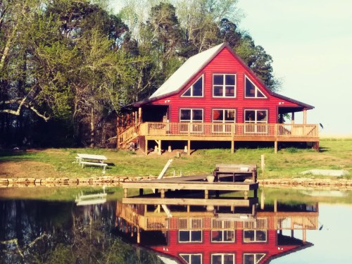 A red cabin with a deck sits by a calm pond, reflecting its image in the water, surrounded by trees.