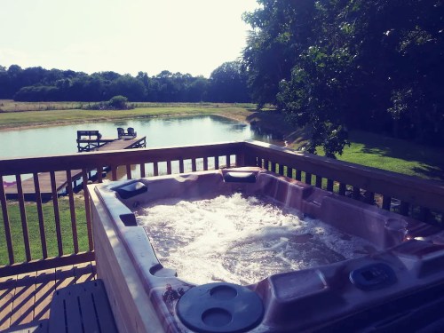 A hot tub on a wooden deck overlooking a serene pond and green landscape under a clear blue sky.