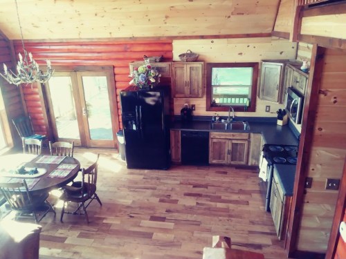 Cozy log cabin kitchen with wooden cabinets, a black fridge, and a dining table, featuring natural light and rustic decor.