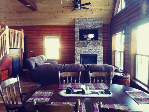 Cozy log cabin interior with a stone fireplace, large windows, and a dining table set for meals.