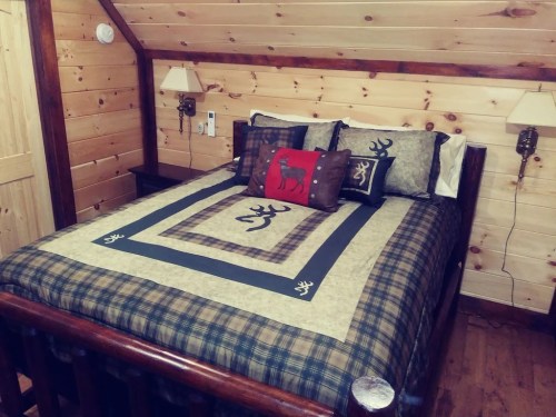 Cozy cabin bedroom with a wooden bed, plaid bedding, and decorative pillows against a rustic wood-paneled wall.