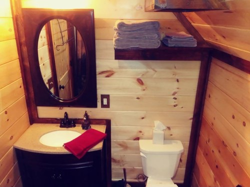 Cozy bathroom with wooden walls, a round mirror, sink, toilet, and neatly stacked towels on a shelf.