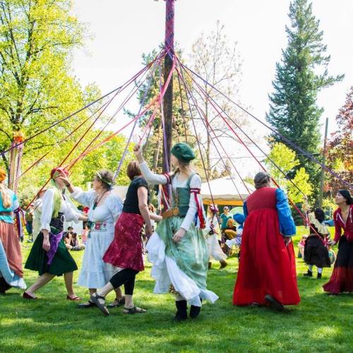 A group of people in colorful costumes dancing around a maypole in a sunny park setting.