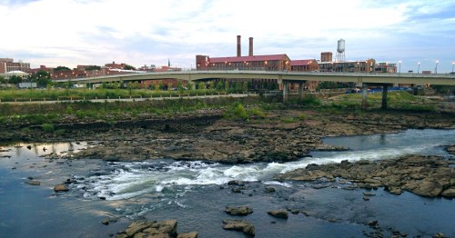 A river flows over rocky terrain with a bridge and industrial buildings in the background under a cloudy sky.
