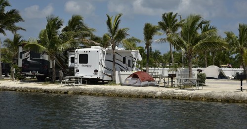 A scenic campsite by the water featuring RVs, tents, and palm trees under a partly cloudy sky.