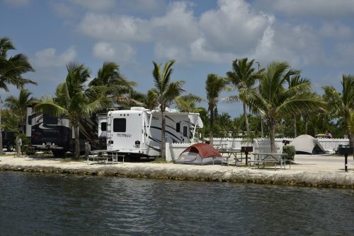 A scenic campsite by the water, featuring RVs, tents, and palm trees under a partly cloudy sky.