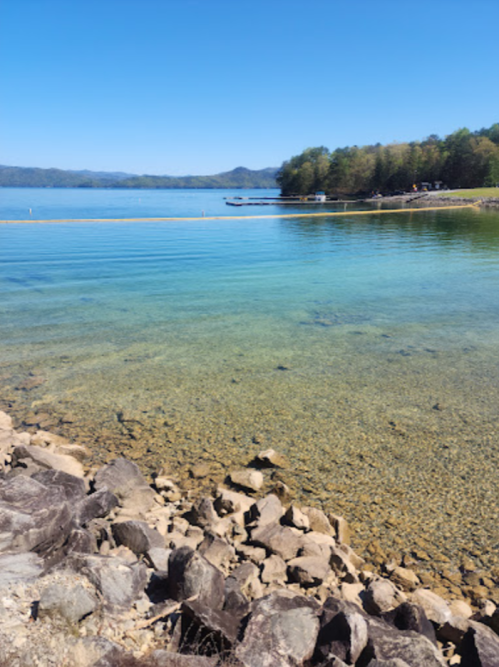 A serene lake with clear water and rocky shoreline, surrounded by green trees under a blue sky.