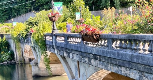 A stone bridge adorned with flower baskets, surrounded by lush greenery and a person walking in the background.