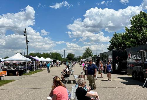 A bustling outdoor market with vendors, food trucks, and visitors enjoying a sunny day under a blue sky with clouds.