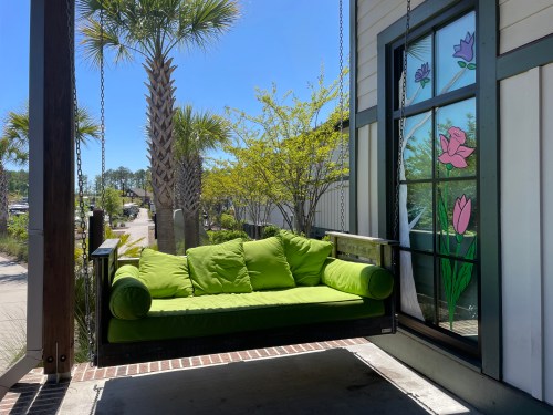 A bright green swing couch on a porch, with palm trees and colorful flower window art in the background.
