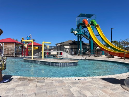 A sunny pool area featuring a water slide, splash features, and a clear blue sky.