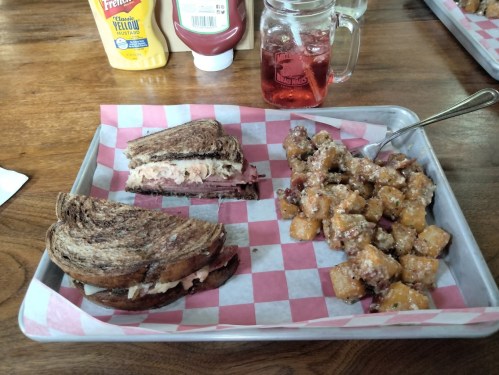 A tray with a sandwich on dark bread, a side of seasoned potato cubes, and a drink in a mason jar.