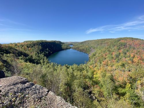 A serene lake surrounded by lush green and autumn-colored trees under a clear blue sky.
