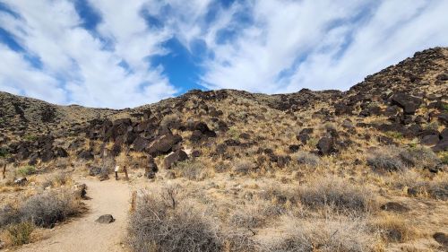 A rocky hillside with sparse vegetation under a blue sky with clouds, featuring a dirt path leading into the landscape.
