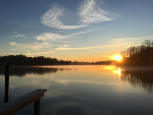 A serene sunrise over a calm lake, with mist rising and trees silhouetted against the colorful sky.