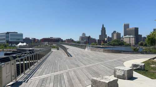 A wooden walkway curves along a river, with city buildings and a clear blue sky in the background.