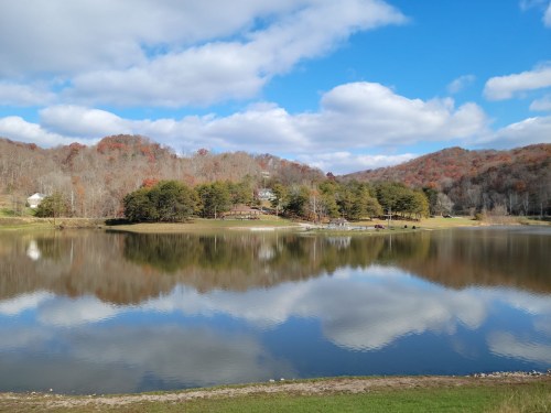 A serene lake reflects trees and clouds, surrounded by autumn-colored hills under a blue sky.