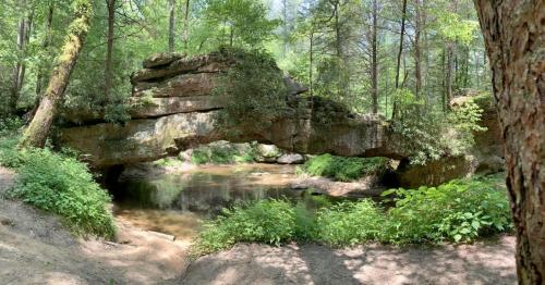 A natural stone arch over a calm stream, surrounded by lush greenery and tall trees in a serene forest setting.