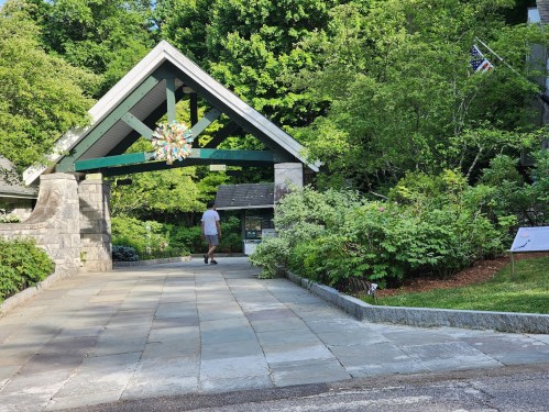 A person walks through a stone archway surrounded by greenery and trees on a sunny day.