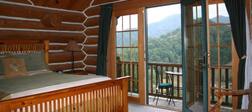 Cozy log cabin bedroom with a wooden bed, large windows, and a view of mountains and greenery outside.