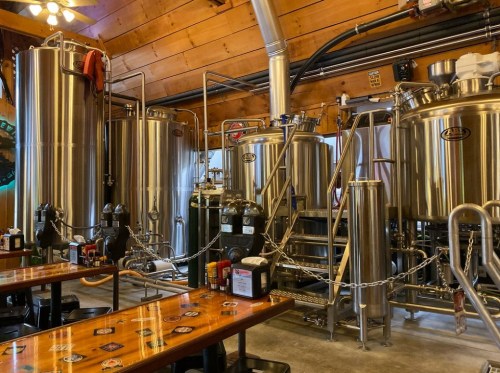 Interior of a brewery featuring stainless steel brewing equipment and wooden tables.