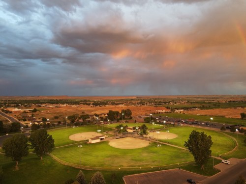 Aerial view of a baseball field at sunset, with dramatic clouds and a rainbow in the background over a rural landscape.