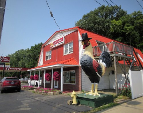 A large chicken statue wearing a hat stands outside a bakery with a red roof and flower planters.
