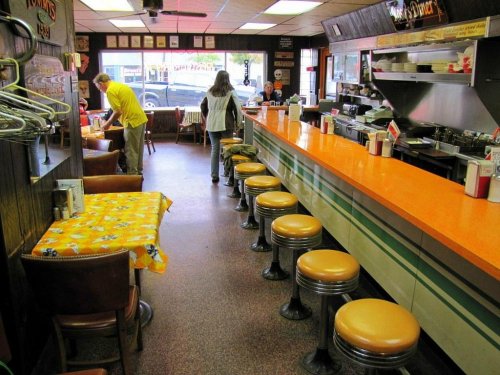 A retro diner interior with orange countertops, metal stools, and tables, featuring customers and a bustling atmosphere.