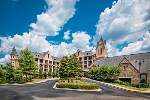 A scenic view of a hotel with stone and brick architecture, surrounded by greenery and a blue sky with fluffy clouds.