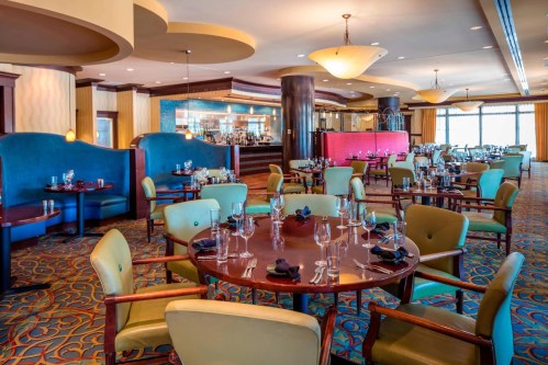 Elegant restaurant interior with round tables, green chairs, and a bar in the background, featuring warm lighting.