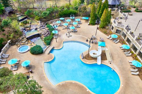 Aerial view of a resort pool area featuring a large pool, slide, hot tub, and lounge chairs under teal umbrellas.